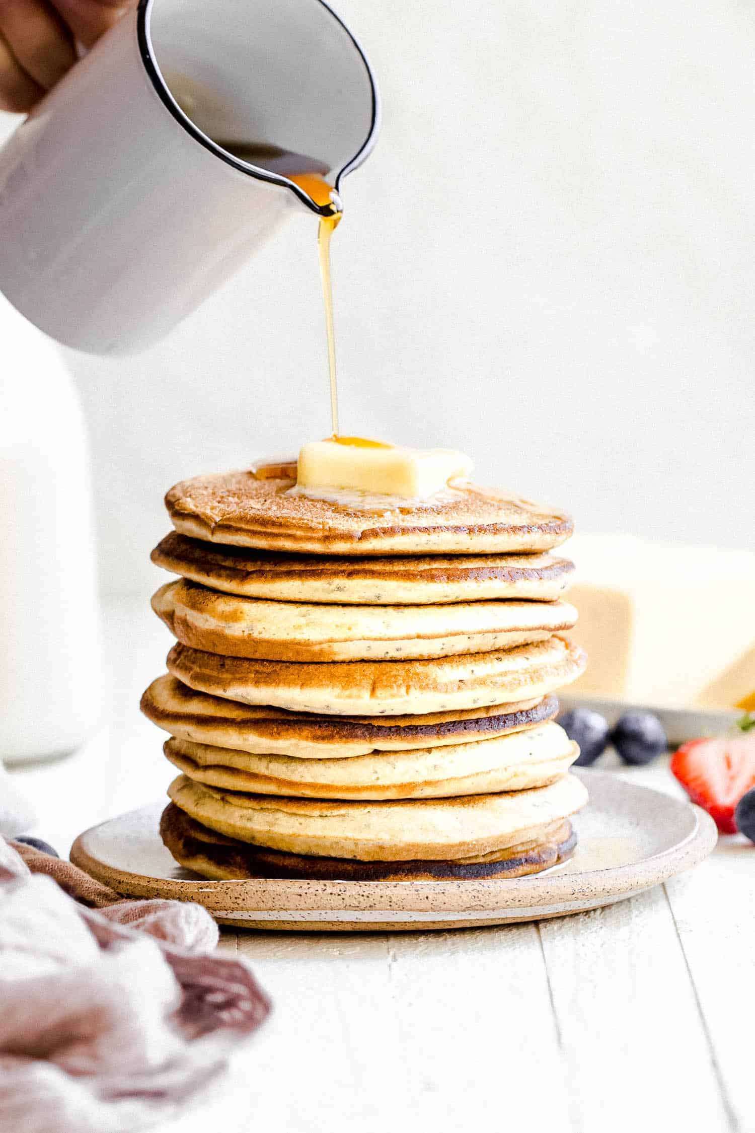 pouring maple syrup onto a stack of fluffy pancakes on a white plate