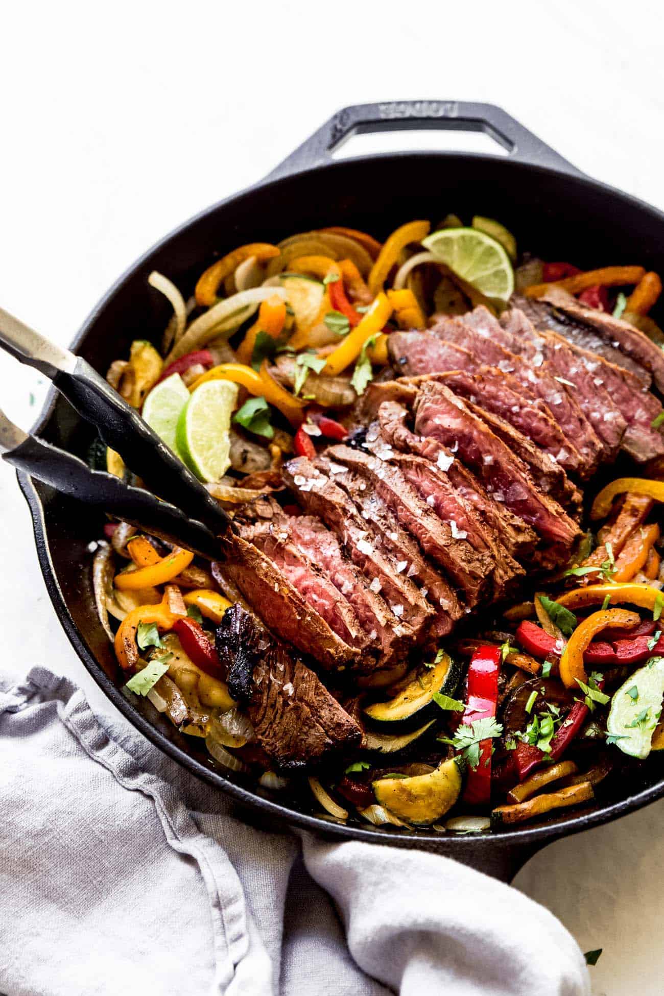 tongs picking up steak fajitas out of a cast iron pan