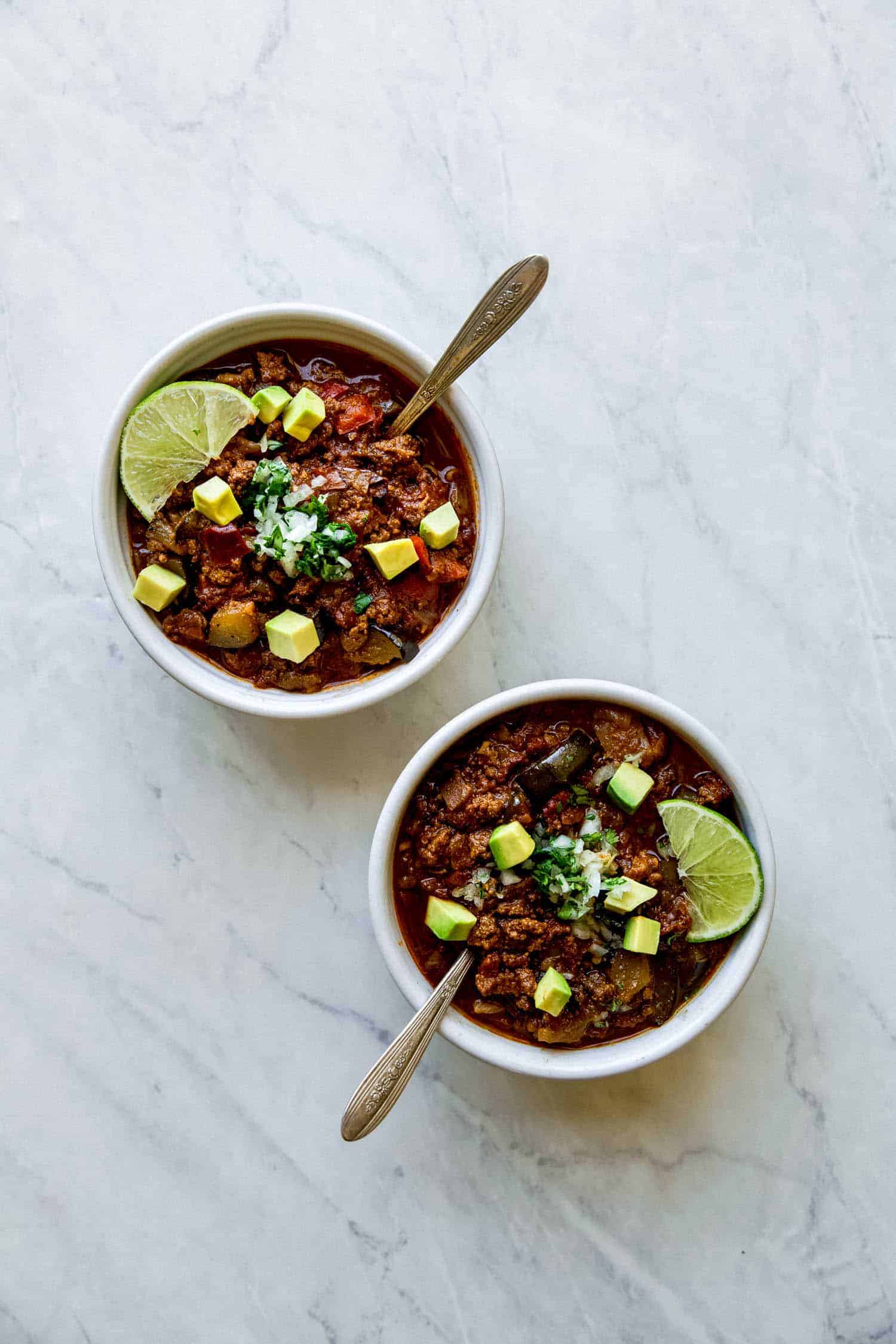 two bowls of paleo chili with spoons inside and avocado chunks and lime on top