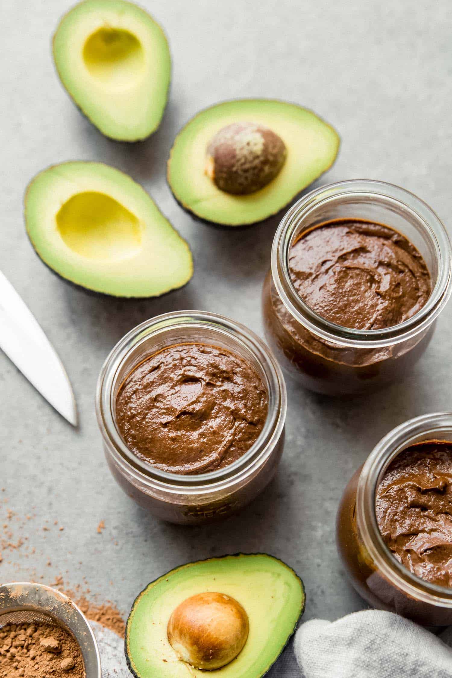 chocolate avocado pudding in jars with avocados on the side on a grey board