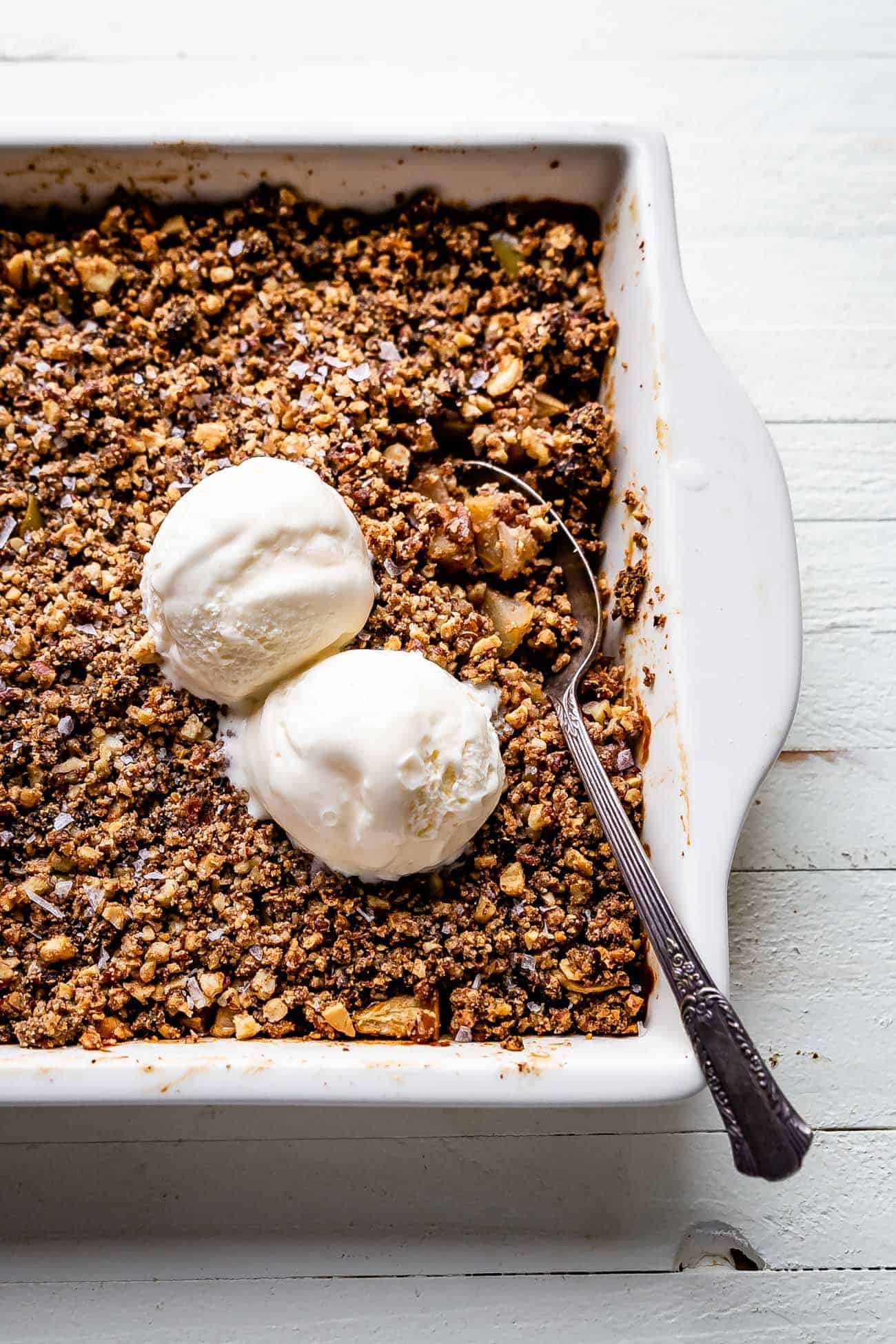 paleo apple crisp in a white dish with a spoon inside and vanilla ice cream