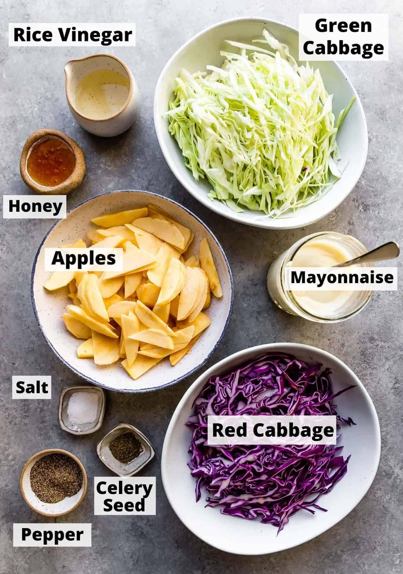 ingredients laid out in bowls to make creamy coleslaw recipe: rice vinegar, green cabbage, honey, mayonnaise, apples, salt, red cabbage, celery seed, pepper