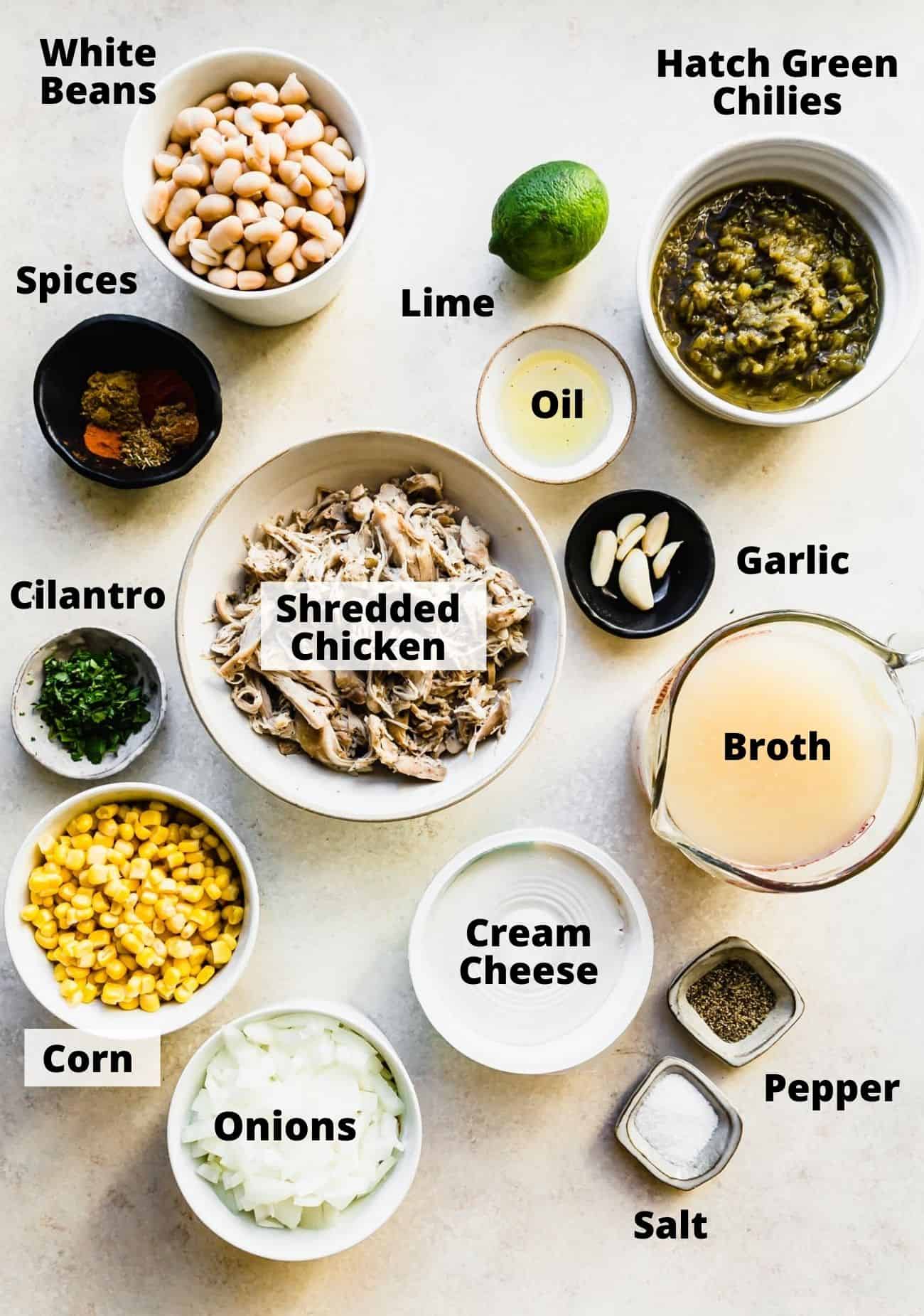 ingredients laid out to make instant pot white chicken chili: white beans, hatch greens chilies, spices, lime, oil, garlic, broth, shredded chicken, cilantro, corn, onions, cream cheese, pepper, salt