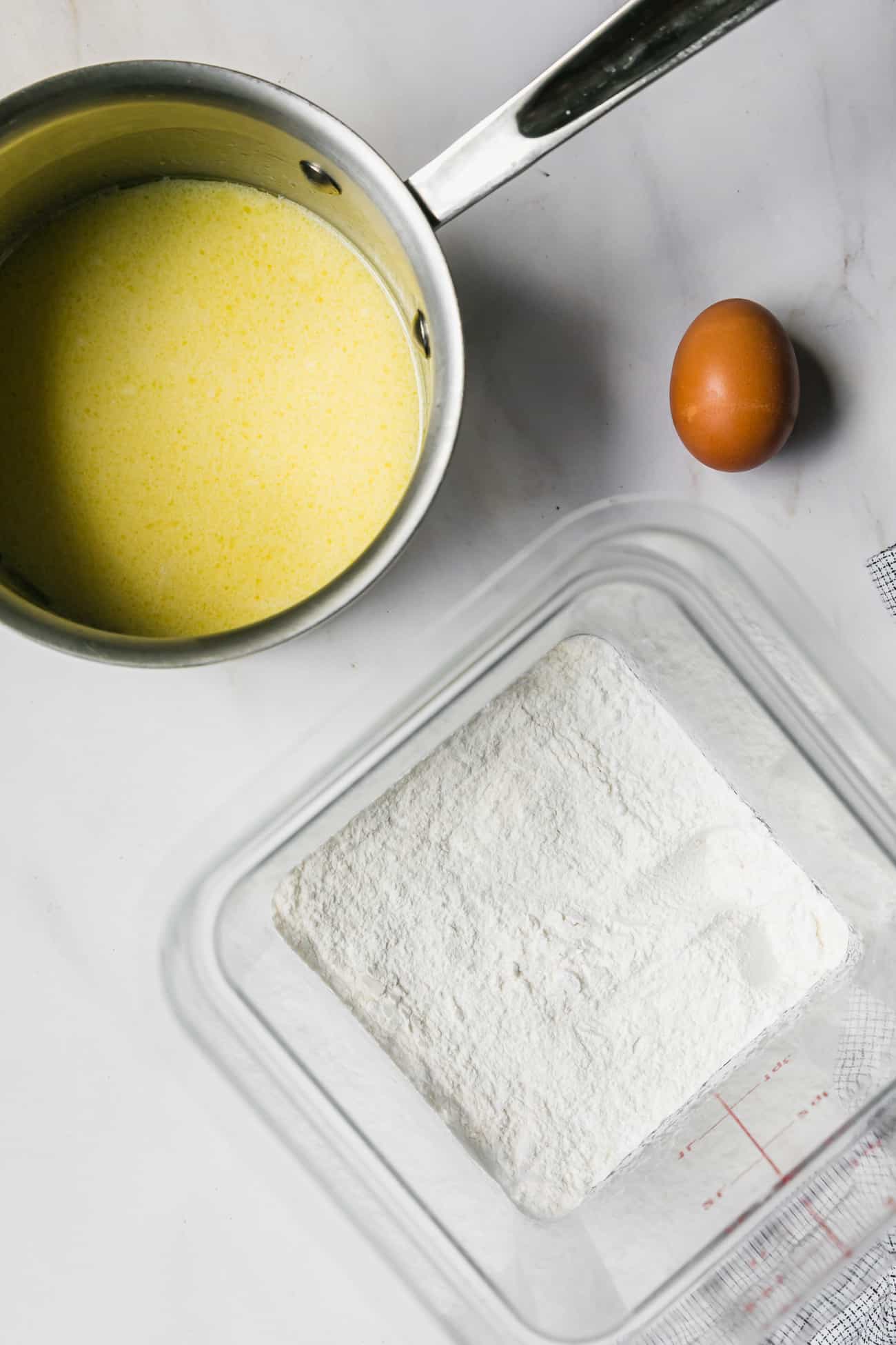 ingredients prepped to make 5 ingredient pizza dough: a saucepan, clear square bin with flour and an egg