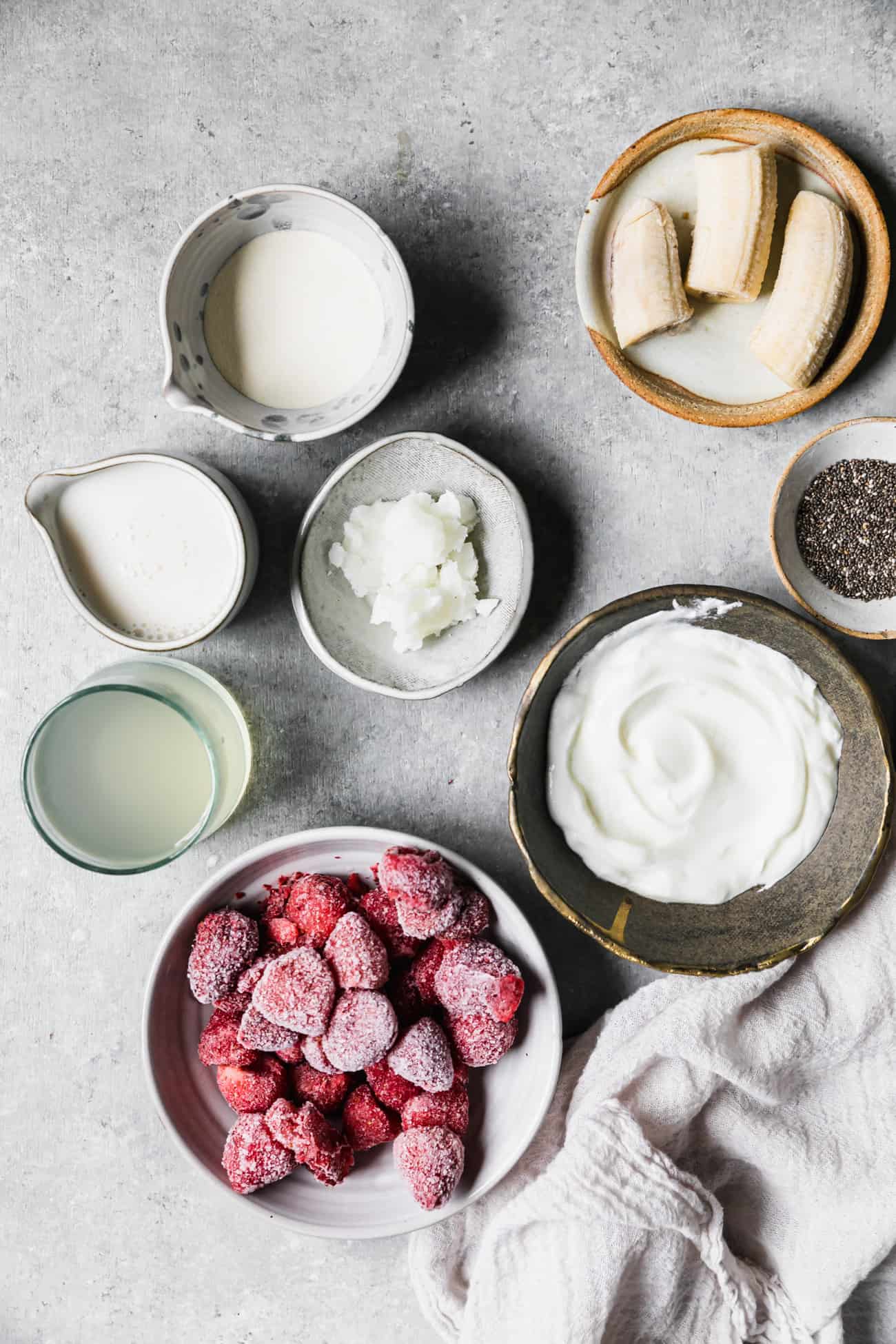 ingredients for yogurt smoothie bowl prepped: frozen strawberries, frozen banana, milk, coconut water, collagen, chia seeds, yogurt, coconut oil.