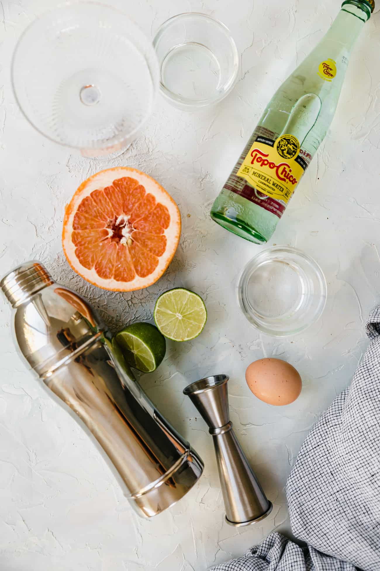 ingredients laid out to make a Paloma: grapefruit, sparkling water, limes, an egg, tequila, and simple syrup.